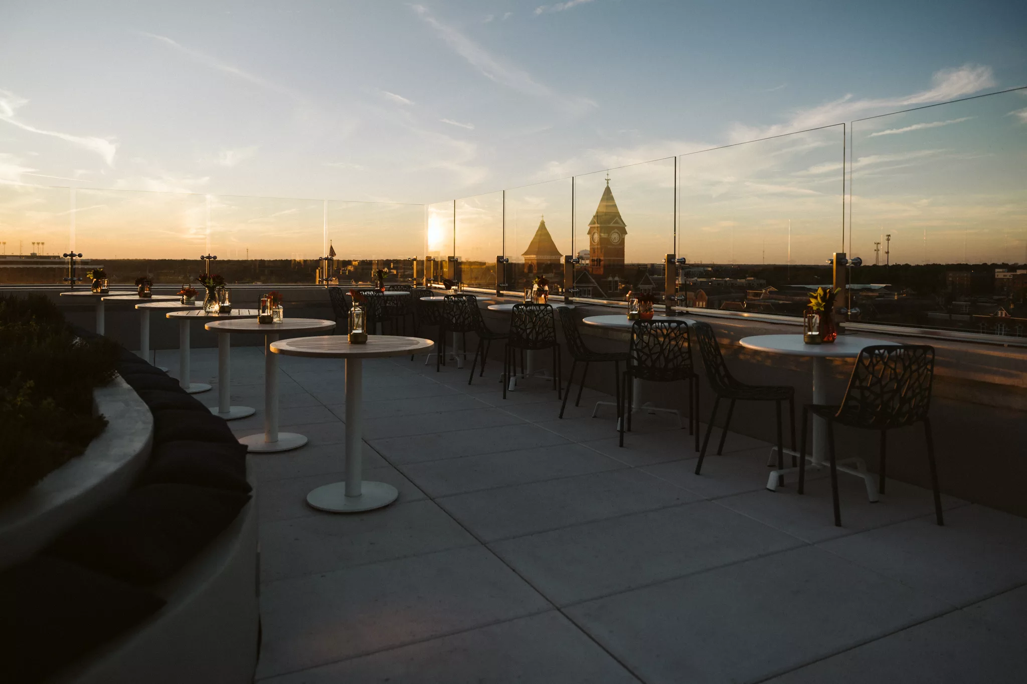 A rooftop terrace at sunset with white tables and dark chairs arranged along the glass barrier. The sky is tinged with orange and blue hues, and there are distant cityscape views with a prominent tower and spire, all contributing to a serene atmosphere. - Hausion