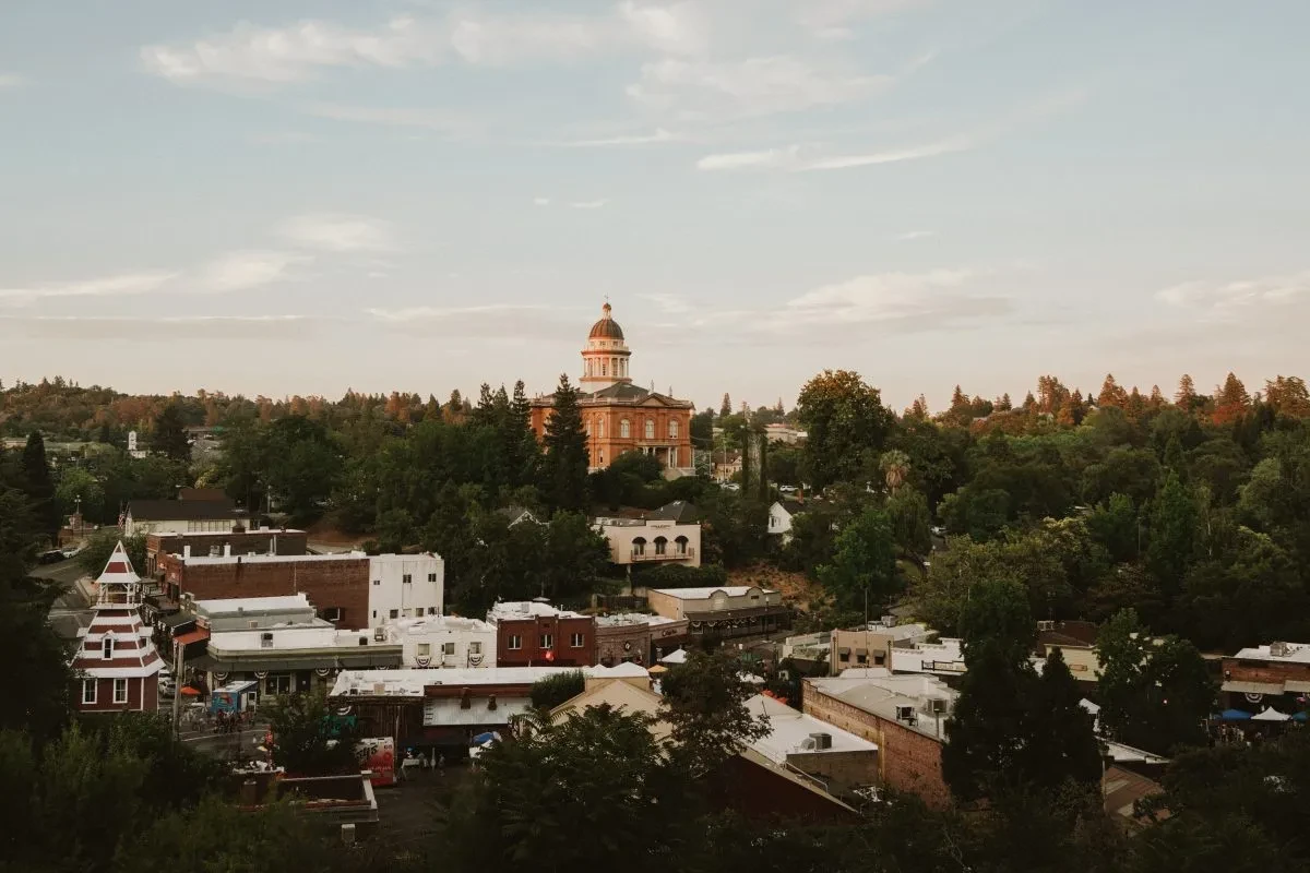 Aerial view of a small town featuring a historic courthouse with a dome at its center, surrounded by trees and old buildings. The weather is clear with a few clouds in the sky, and the scene has a peaceful, rural atmosphere. - Hausion