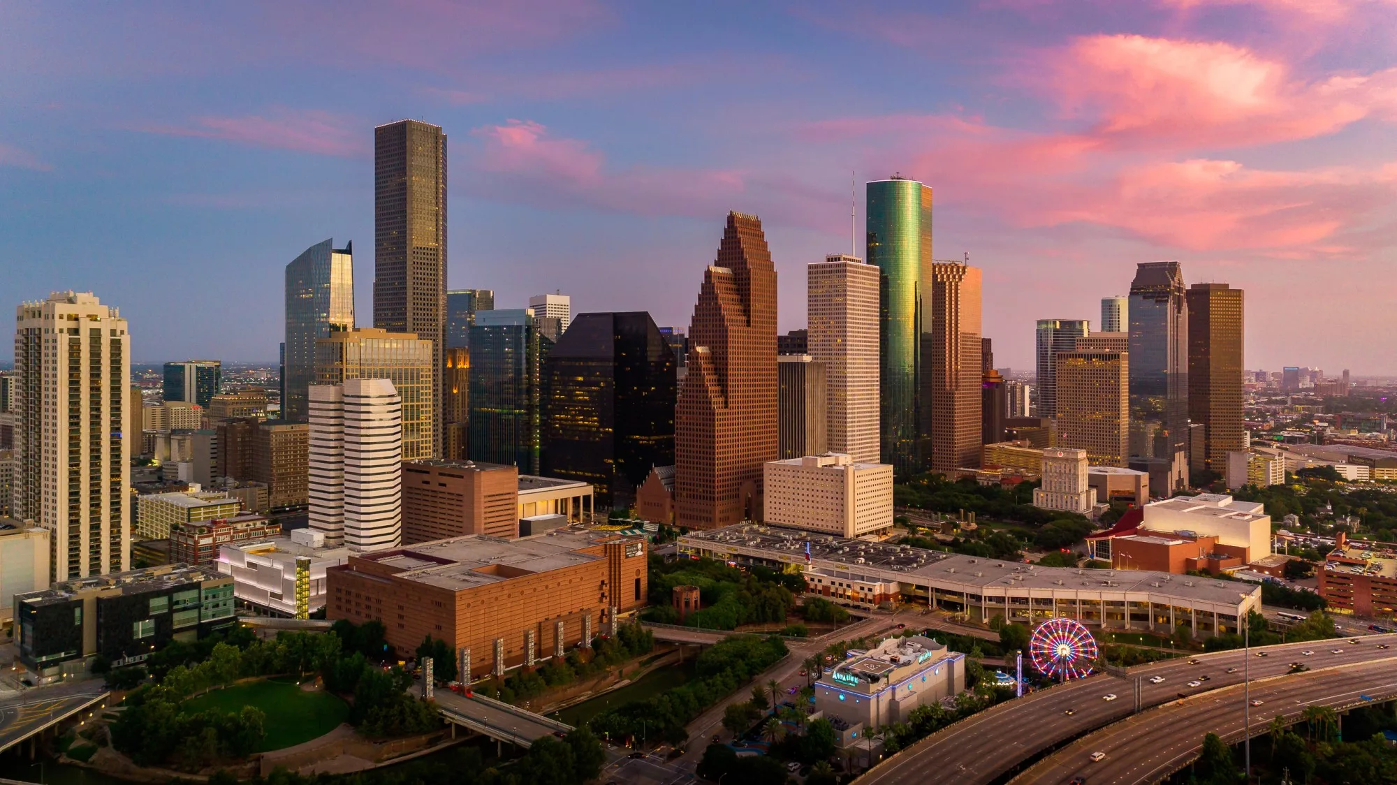 Aerial view of a cityscape during sunset, showcasing modern high-rise buildings with unique architectural designs. The sky is adorned with pink and purple hues. A Ferris wheel and a busy highway are visible in the foreground, adding vibrancy to the urban scene. - Hausion